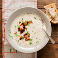 A bowl of baked potato soup topped with bacon bits and chives, served with pieces of bread on the side.