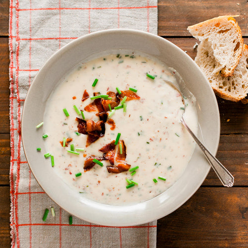 A bowl of baked potato soup topped with bacon bits and chives, served with pieces of bread on the side.