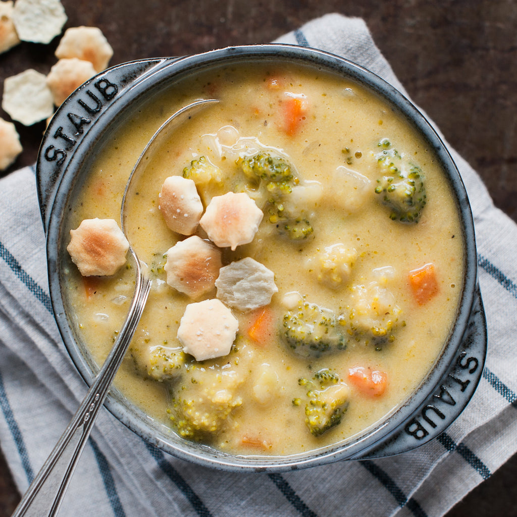 A bowl of broccoli cheddar soup with pieces of broccoli, carrots, and onions visible, garnished with cheese crackers and a spoon.