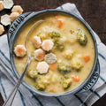 A bowl of broccoli cheddar soup with pieces of broccoli, carrots, and onions visible, garnished with cheese crackers and a spoon.