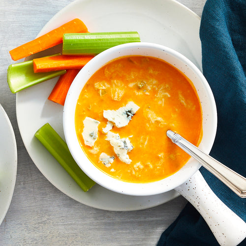 A bowl of Buffalo Chicken Bisque soup with pieces of blue cheese and a spoon, served with celery and carrot sticks on the side, placed on a white plate with a blue napkin.