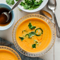 A bowl of carrot and ginger soup garnished with green onions, with a side salad and utensils on the side.
