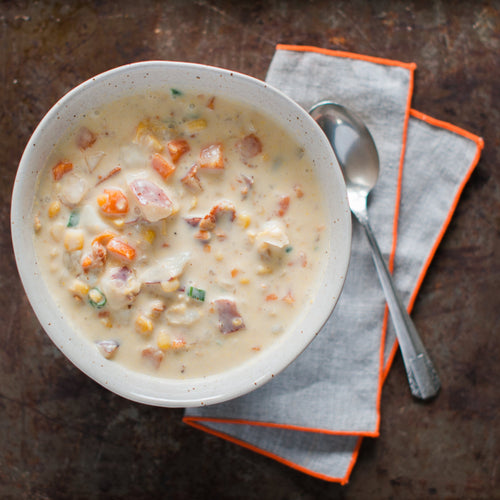 A bowl of Cheddar Corn Chowder soup with visible chunks of corn, potatoes, carrots, and bacon, served on a grey napkin with orange trim.