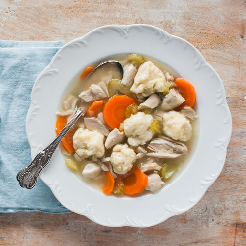 A bowl of Chicken & Dumplings soup with visible ingredients including carrots, celery, onions, and dumplings, served on a white plate with a spoon on the side.
