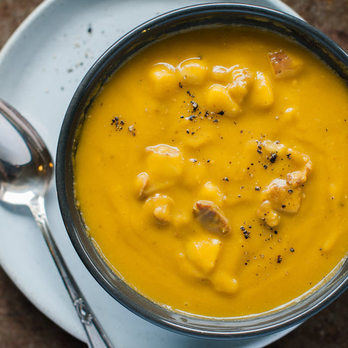 A bowl of Harvest Split Pea soup with visible chunks of potato and bacon, garnished with black pepper.