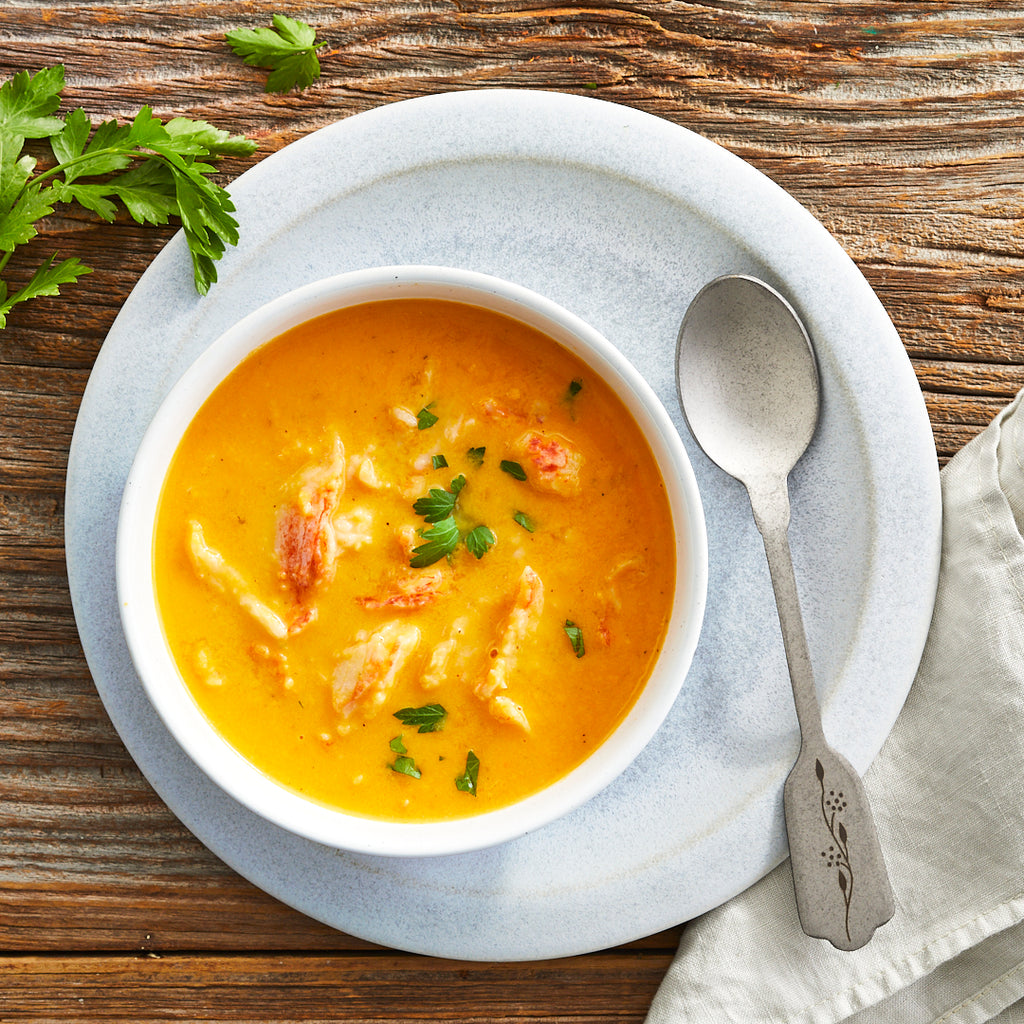 A bowl of lobster bisque soup garnished with parsley, with a spoon on the side, on a wooden table.