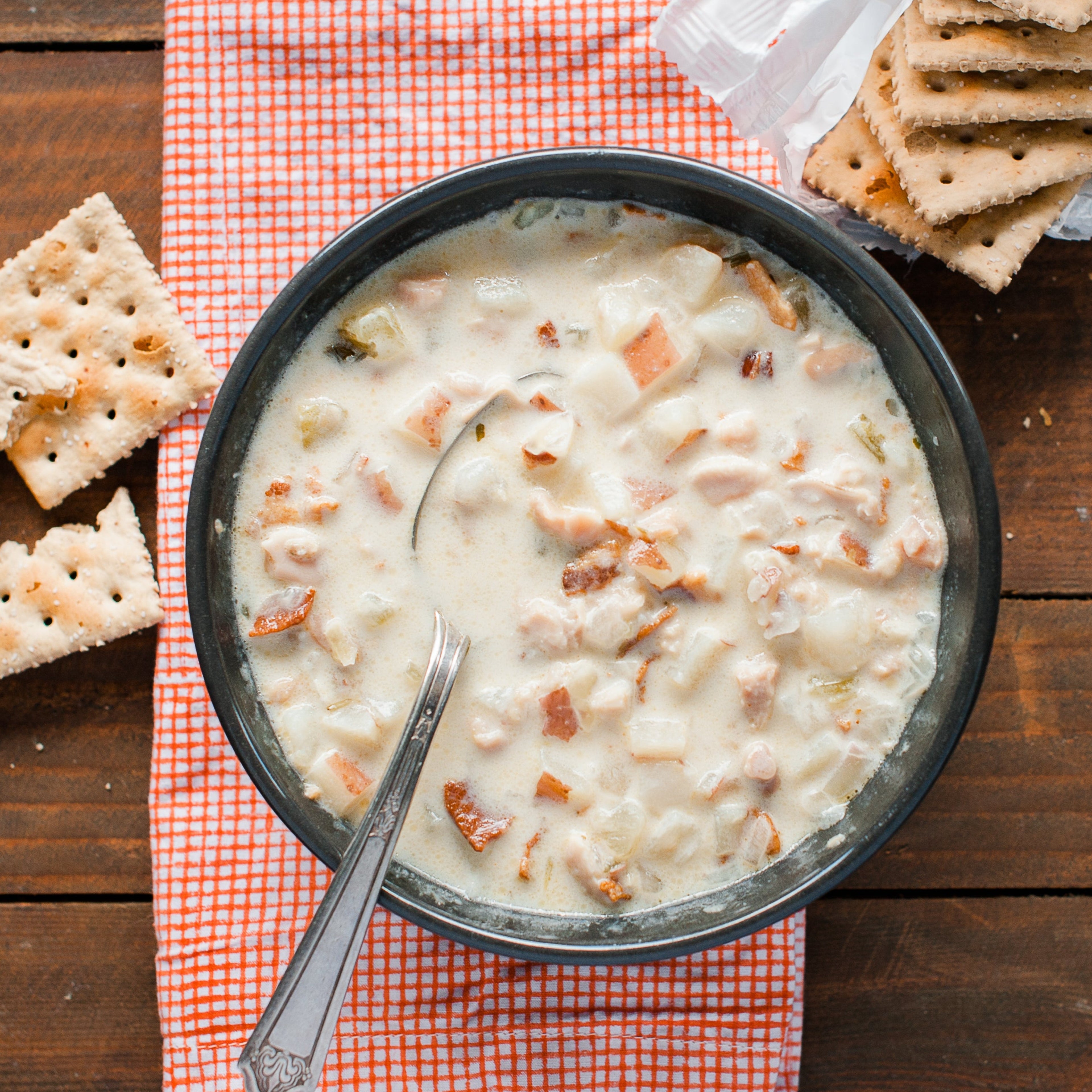 A bowl of New England Clam Chowder with pieces of bacon, onions, and potatoes visible, served on an orange and white checkered napkin with crackers on the side.