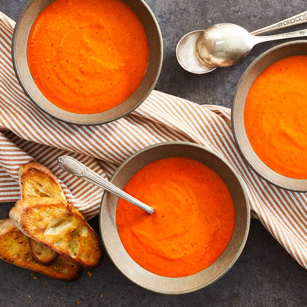 Bowl of red pepper gouda soup with a side of bread and a spoon, on a striped napkin.