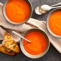 Bowl of red pepper gouda soup with a side of bread and a spoon, on a striped napkin.