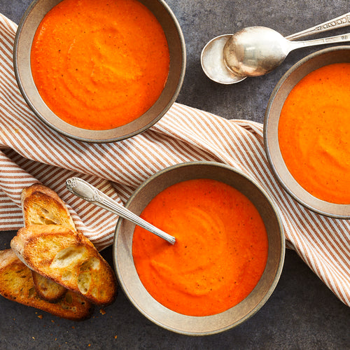 Bowl of red pepper gouda soup with a side of bread and a spoon, on a striped napkin.