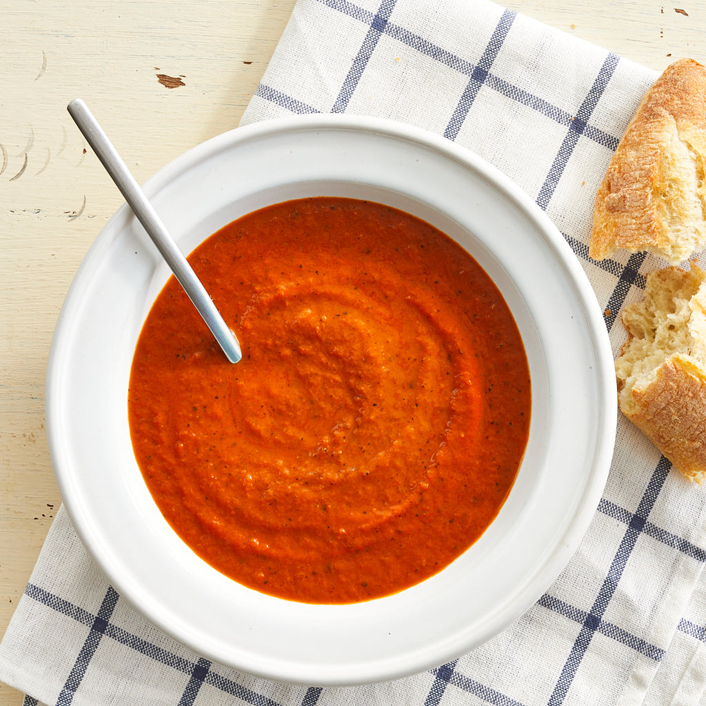 A bowl of roasted tomato bisque soup with a spoon, accompanied by a piece of bread, placed on a white plate with blue stripes.