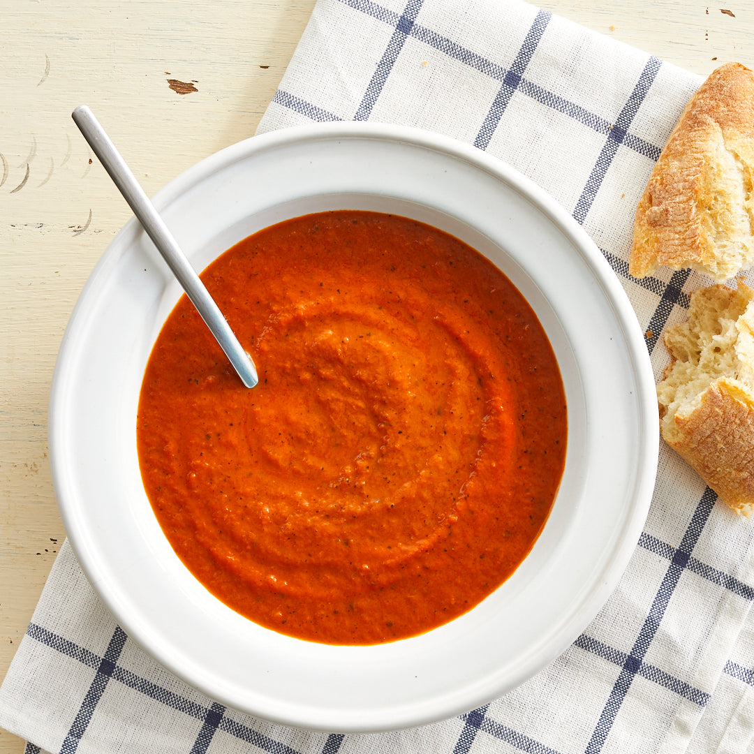 A bowl of roasted tomato bisque soup with a spoon, accompanied by a piece of bread, placed on a white plate with blue stripes.