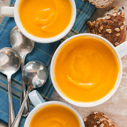 Bowls of root vegetable soup served with bread and spoons on the side, presented on a table with a blue checkered napkin.