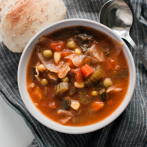 A bowl of vegetable soup containing carrots, peas, corn, and other vegetables, served alongside a roll.