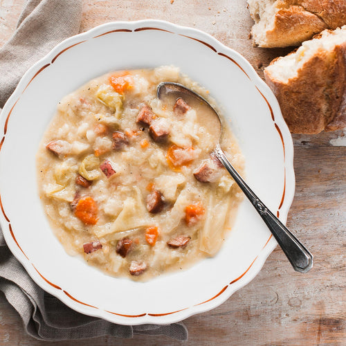 A bowl of Smoked Kielbasa & Cabbage soup with a spoon, alongside pieces of bread, on a wooden surface.
