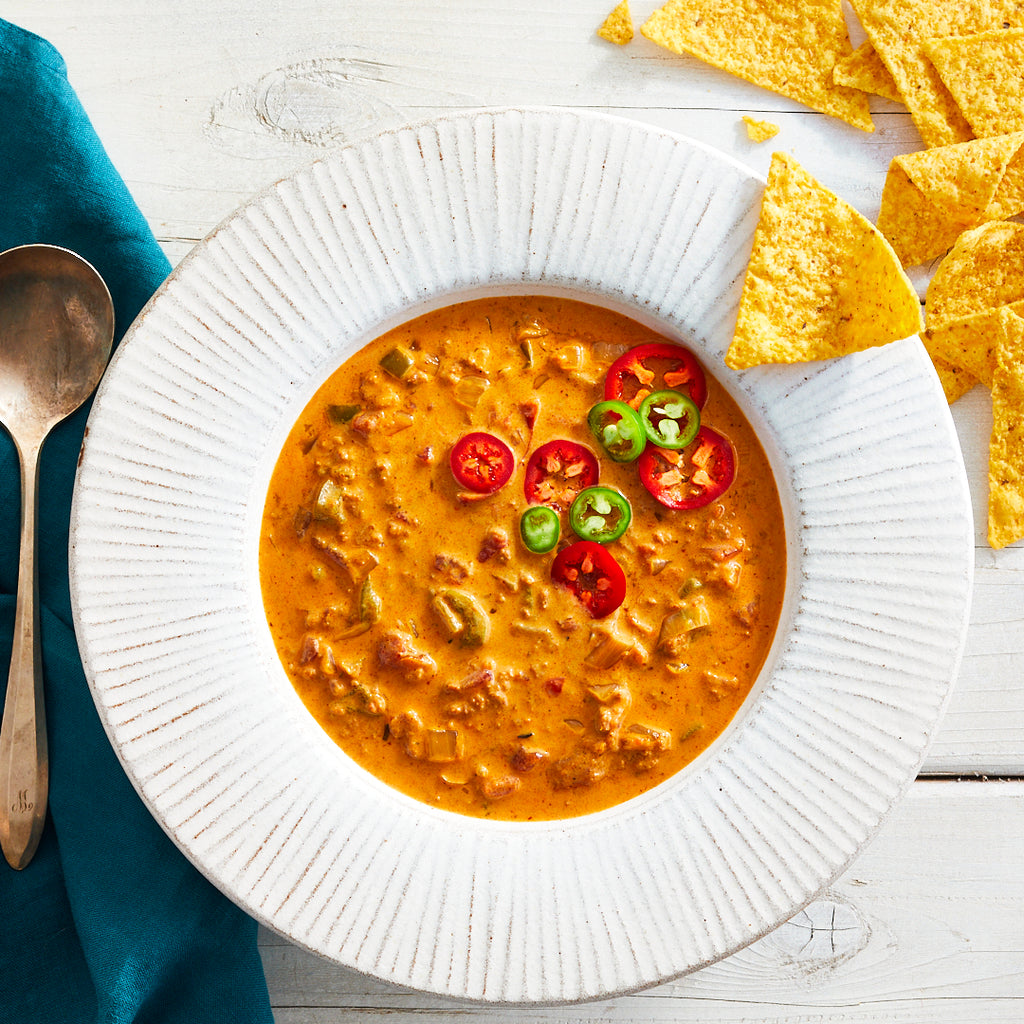 A bowl of spicy chorizo loco soup garnished with sliced jalapeno peppers and tomatoes, accompanied by tortilla chips and a spoon on the side.