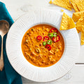A bowl of spicy chorizo loco soup garnished with sliced jalapeno peppers and tomatoes, accompanied by tortilla chips and a spoon on the side.