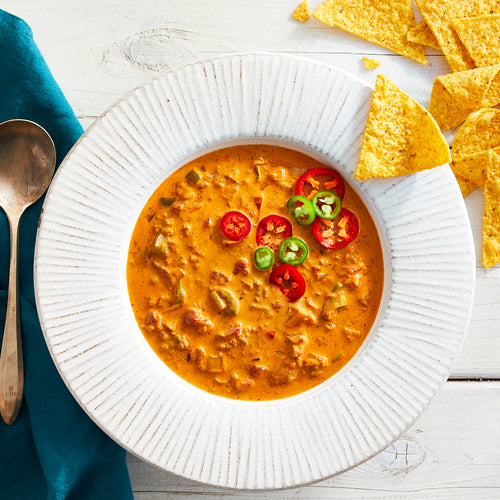 A bowl of spicy chorizo loco soup garnished with sliced jalapeno peppers and tomatoes, accompanied by tortilla chips and a spoon on the side.