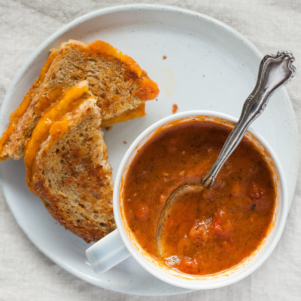 A bowl of tomato and dill soup with a side of grilled cheese sandwich, viewed from above with a spoon in the soup.